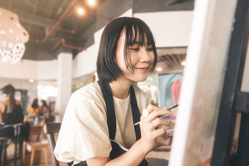 Portrait of happy young adult asian woman painting brush on canvas during online art class.