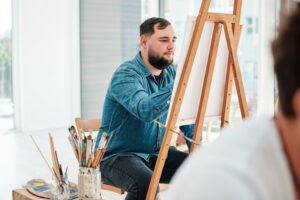 A handsome young artist sitting and painting during an online art class for adults in his studio