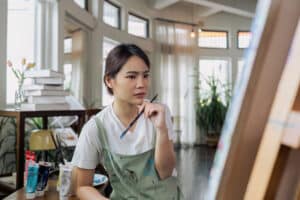 Female artist thinking and hold paintbrush while looking at canvas and pondering about how to fix an acrylic painting mistake. Yarnell School Online - YSO