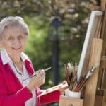 A smiling senior woman is in an outdoor setting painting. Never Too Old to Start Painting - Yarnell School of Fine Art