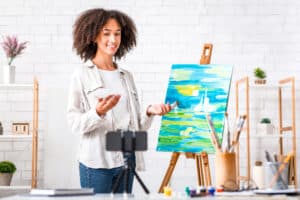 Paid Art Lessons Online. Smiling African American woman with palette and paintbrush stands near easel and looks at webcam at smartphone, at home