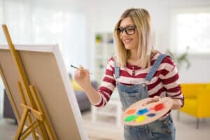 Young female artist painting on a canvas with a paintbrush and holding a color palette at home. acrylic vs. watercolor painting, the joy of experimentation - Yarnell School Online