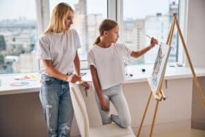 Side view of a female standing behind a girl painting a picture on the canvas. Best painting medium for beginners - Yarnell School of Fine Art